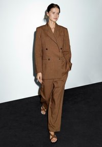 Vrouw die op een zwarte vloer tegen een witte muur loopt, gekleed in een oversized bruine dubbelbreasted blazer en bijpassende wijde broek met bruine sandalen.