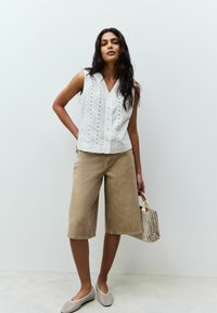 Top sans manches blanc avec détails de bouton et ouvertures en œillet, associé à un short large beige clair et des chaussures à enfiler à motifs.