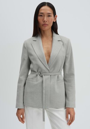 Donna con capelli lunghi e scuri che indossa un blazer grigio chiaro con cintura e pantaloni bianchi, in piedi davanti a uno sfondo grigio uniforme.