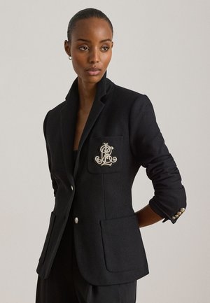 Γυναίκα που φοράει προσαρμοσμένο μαύρο σακάκι με κεντημένο έμβλημα στην τσέπη στο στήθος και γυρισμένα μανίκια, στέκεται μπροστά σε απλό φόντο.