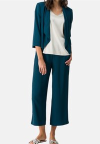 Türkisfarbener Cardigan über einer weißen Bluse, kombiniert mit türkisfarbenen weit geschnittenen Hosen. Das Material wirkt weich. Akzentuiert durch ein leichtes Design. Glänzende Sandalen.