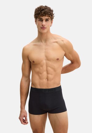 Spierige man met krullend haar, gekleed in zwarte Marc O'Polo boxer briefs, staand tegen een eenvoudige lichte achtergrond met één arm achter zijn rug.