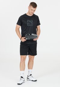 Svart tränings t-shirt med "PAD" och "EL" tryckt i grått, ihop med svarta shorts. Individuen håller en padelracket och har på sig vita träningsskor.