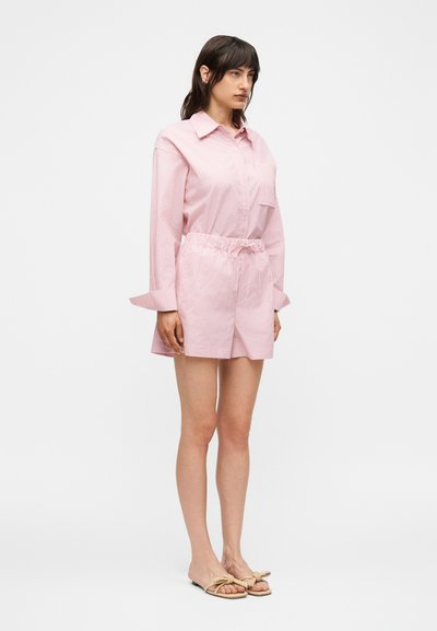 Γυναίκα που φοράει ροζ μπλούζα με μακριά μανίκια και ταιριαστά σορτς με μπεζ σανδάλια slide με κόμπο, σε απλό λευκό φόντο.