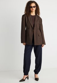 Γυναίκα με κατσαρά μαλλιά που φοράει σκούρα γυαλιά ηλίου, καφέ υπερμεγέθη σακάκι, μπλε παντελόνι τύπου τζόγκερ και μαύρα ψηλοτάκουνα παπούτσια με λουράκι, στέκεται όρθια.