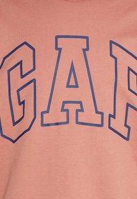 Sudadera de algodón rosa con grandes letras azules "GAP". El material tiene una textura suave y un diseño de cuello redondo.