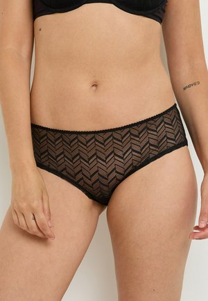 Donna che indossa perizoma nero con un motivo in pizzo e reggiseno nero, mostrando il ventre e parzialmente le braccia con tatuaggi sul braccio destro e sulla mano sinistra.