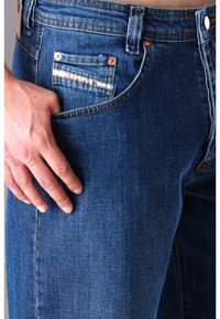 Jeans aus Denim in einem tiefblauen Farbton, mit kupferfarbenen Akzenten und einer dekorativen kleinen Tasche mit einem weißen horizontalen Streifen.
