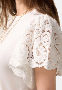 Top bianco a maniche corte con dettagli in pizzo elaborati sulle spalle, caratterizzato da motivi floreali e spirali, in tessuto morbido e con una vestibilità comoda.