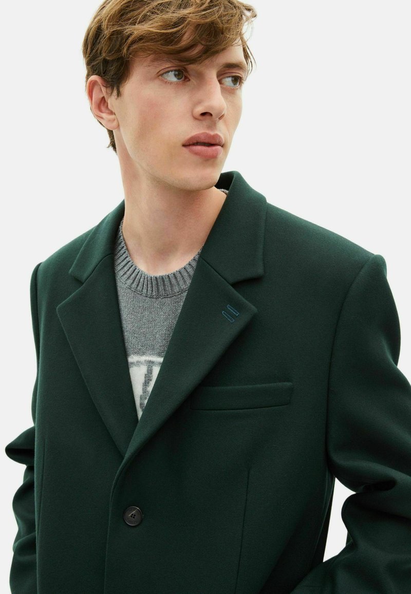 Dark green wool blazer with a notched collar, single button closure, and a chest pocket; worn over a light gray knitted sweater.