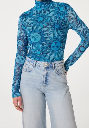 Blaues durchsichtiges Langarmoberteil mit Sonnen- und Herzmustern, kombiniert mit hellblauen Jeans mit hoher Taille und einem goldenen Ring an der linken Hand.