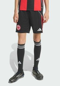Svarta fotbollsshorts med röda detaljer och ett emblem, kombinerat med svarta strumpor med vita ränder och svarta fotbollsskor med grå detaljer.