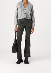 Grijze gebreide cardigan met kabelpatroon, V-hals en knopen, gecombineerd met olijfgroene uitlopende ribfluwelen broek en zwarte enkellaarsjes, met een zwarte tas in de hand.