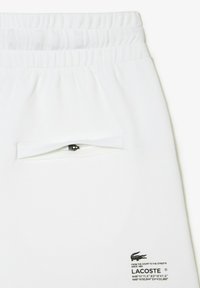 Pantaloni corti bianchi da allenamento con una vita elastica, dotati di una piccola tasca con zip e un logo nero Lacoste in basso a sinistra.