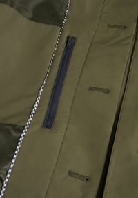 Olívazöld dzseki belseje fényes textúrájú, fekete cipzáras zsebbe és sakktáblas varrásba öltözött az élein.