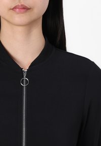 Una donna con una giacca nera con un inserto a zip argentato, che mostra il collo e le labbra su uno sfondo bianco.