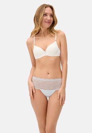 Mujer con sujetador acolchado blanco y ropa interior a juego de encaje, de pie contra un fondo gris claro, mirando hacia un lado con una ligera sonrisa.