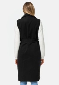 Schwarzer ärmelloser Mantel mit hohem Kragen, tailliertem Schnitt und weichen weißen Ärmeln. Verfügt über einen strukturierten Stoff und ein elegantes Design.