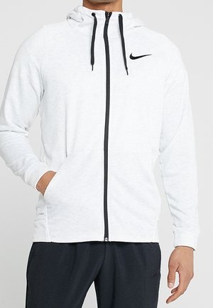 Mann trägt einen weißen Nike-Hoodie mit Reißverschluss, schwarzen Kordeln und Logo, kombiniert mit schwarzen Hosen, Hände in den Taschen und entspannte Pose.