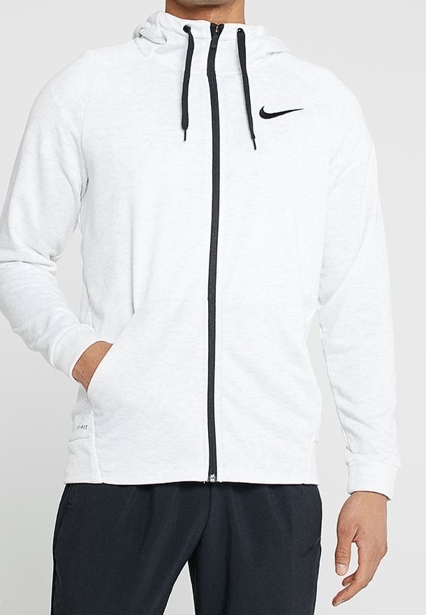Homme portant un sweat à capuche Nike blanc zippé avec des cordons noirs et un logo, associé à un pantalon noir, mains dans les poches et posture détendue.