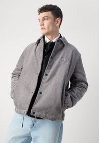 Grå cordjakke med knappelukking, med krage og rillestruktur, parret med en svart skjorte og lyseblå jeans.