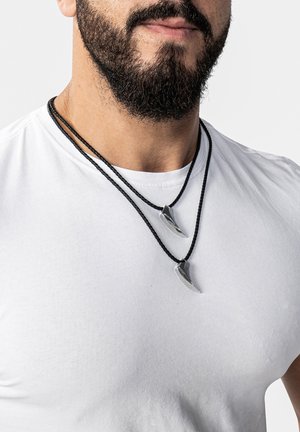 Silberfarbene Krallen-Anhänger an zwei schwarzen geflochtenen Kordeln, getragen über einem schlichten weißen T-Shirt. Strukturierte Oberflächen und geschwungene Formen betonen das Design.