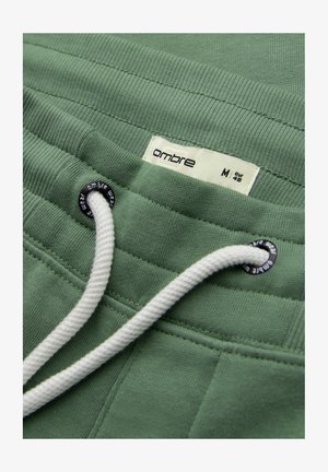 Groene tailleband van een joggingbroek met een wit trekkoord door zwarte metalen oogjes, met het label "ombre" en het maatlabel M EUR 48.