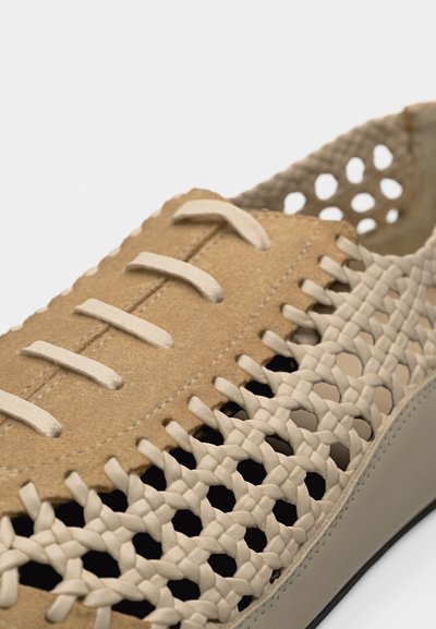 Gros plan sur une chaussure en daim beige avec des côtés en cuir tressé beige et des lacets assortis sur fond blanc.