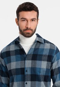 Camisa de franela a cuadros azul y gris con un bolsillo en el pecho, combinada con un cuello alto blanco. Presenta un cuello y botones en la parte frontal.