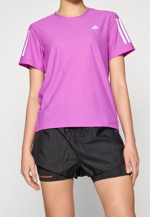 Persona con una camiseta deportiva morada brillante con rayas blancas en las mangas y pantalones cortos de correr negros, de pie frente a un fondo liso.