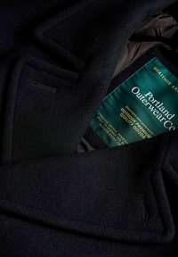 Jūras spēku zila vilnas mētelis ar izdobītu apmali, teksturētu audumu un Portland Outerwear Co. etiķeti ar zaļiem un zeltītiem burtiem.