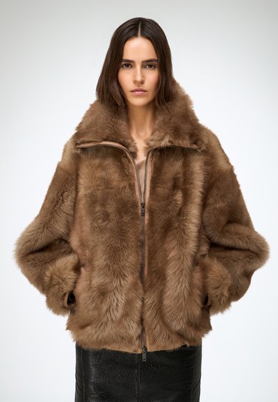 Bruine bontjas met een hoge kraag, ritssluiting aan de voorkant en oversized mouwen. Zachte textuur met een fluweelachtige afwerking. Draagt over een zwarte leren rok.