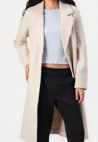 Beige ullkappa med skräddarsydd design, urklippta slag och sidofickor, bärs öppen över en ljusblå texturerad topp och svarta byxor.