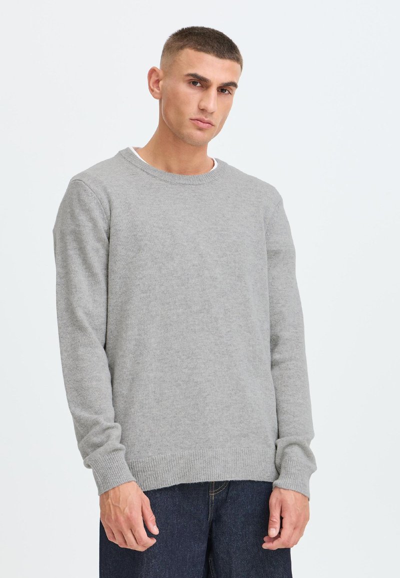 Mand med kort hår iført en lysegrå langærmet sweater og mørkeblå jeans, stående foran en ensfarvet hvid baggrund.