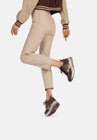 Jeans beige a effetto texture con una vestibilità slim, abbinati a sneakers marroni scure con un design gradient e accenti metallici.