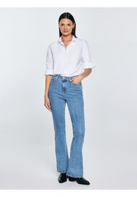 Žena stojící, oblečena v bílé košili na knoflíky s vyhrnutými rukávy, světle modré džíny s vysokým pasem a rozšířenými nohavicemi a černé boty na podpatku.