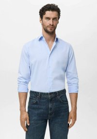 Man met kort, krullend haar in een lichtblauwe overhemd met opgerolde mouwen en donkerblauwe jeans tegen een effen achtergrond.
