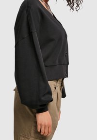 Black cropped cardigan with a v-neck, button closure, and oversized sleeves. Made of soft fabric with ribbed cuffs. Styled with beige pants.