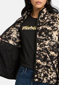 Fleecejacka med ett svart och beige abstrakt mönster, dragkedja fram och två sidofickor. Bärs över en svart t-shirt med grön text.