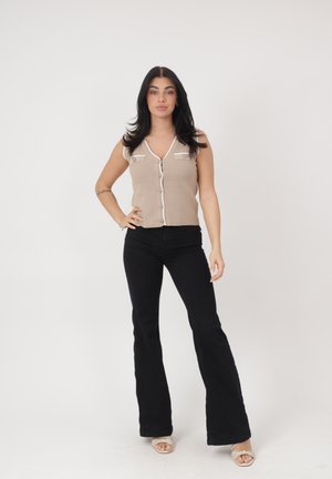 Vrouw met lang donker haar staat met hand op heup, draagt een beige mouwloze top met knopen, zwarte uitlopende broek en beige sandalen.