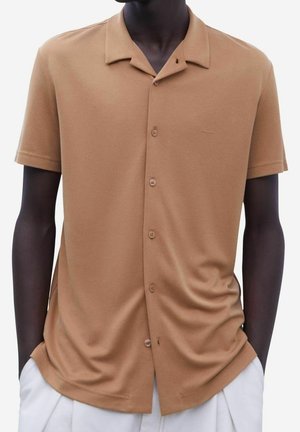 Personne à la peau foncée portant une chemise marron clair à manches courtes avec un col détendu et un pantalon blanc, les mains dans les poches.