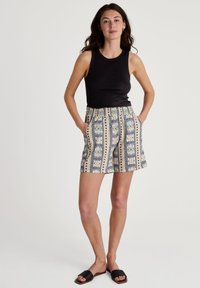 Zwarte mouwloze top, patroonshorts met blauwe, groene en gele ontwerpen, elastische tailleband, zakken, gecombineerd met zwarte schuifslippers.