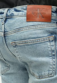 Džínsy Calvin Klein vo svetlom modrom denime majú hnedý kožený pásik s vytlačeným logom a dve zadné vrecká so zvýrazneným šitím.