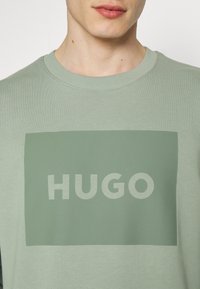 Sudadera verde claro con un diseño rectangular que contiene la palabra "HUGO" en una fuente audaz de un verde ligeramente más claro. Material de algodón.