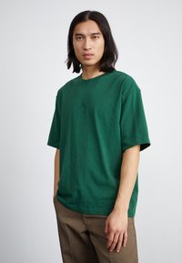 Grøn bomulds T-shirt med rund hals og korte ærmer, der har et subtilt logo på brystet. Båret med lysebrune bukser.