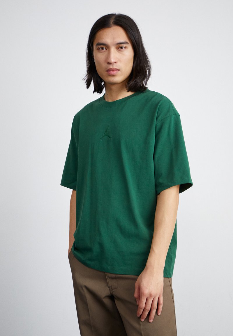 Grøn bomulds T-shirt med rund hals og korte ærmer, der har et subtilt logo på brystet. Båret med lysebrune bukser.