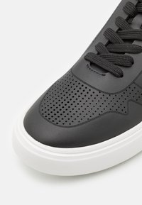 Zapatillas de cuero negro con diseño perforado en la puntera, cordones redondos negros y una suela de goma blanca. Textura suave en todo su diseño.