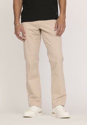 Homme portant un pantalon beige, des baskets blanches, un tee-shirt noir, un bracelet argenté au poignet gauche et une bague argentée au doigt du milieu gauche.