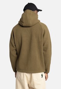 Brun fleece hættebluse med en struktureret finish, der har en stor hætte og en afslappet pasform, parret med lyse bukser med kaktusbroderi.