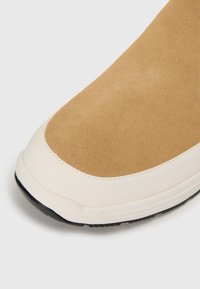 Parte superior de ante marrón con suela de goma crema, que presenta una textura suave y una puntera redondeada con costura a lo largo de la costura.